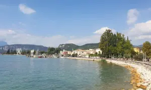 Il lungolago di Bardolino, con il lago azzurro, le barche, le case colorate e le montagne sullo sfondo. Una ruota panoramica si staglia all'orizzonte.