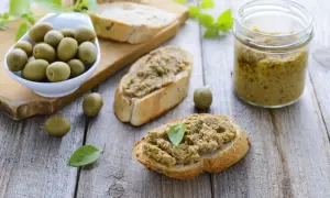 Bruschette con crema di olive, un vasetto di crema di olive e olive verdi su un tagliere di legno.