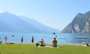 Gente in spiaggia e in acqua, con montagne sullo sfondo, in una giornata di sole sul lago di Garda, forse vicino a Lazise o Bardolino.