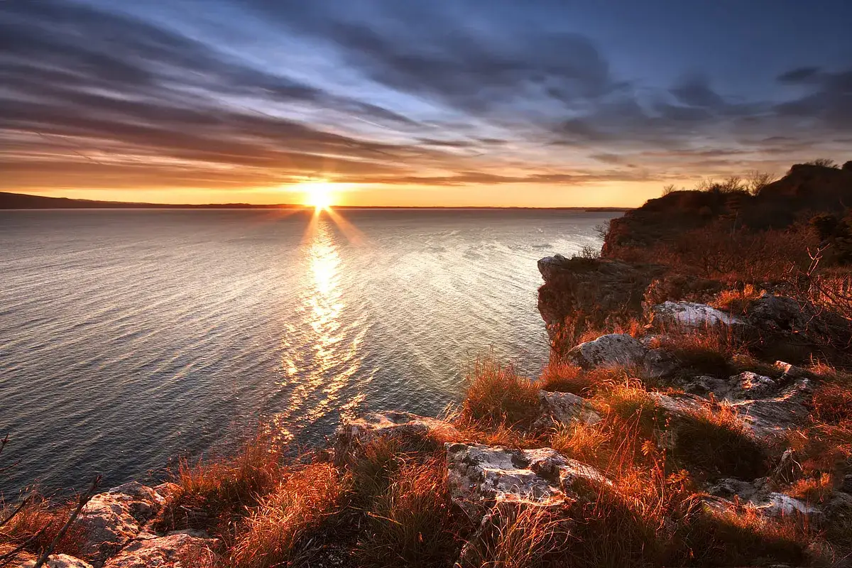 Tramonto dorato sul lago, con scogliere rocciose e vegetazione secca illuminate dal sole. Vista mozzafiato da Manerba del Garda.