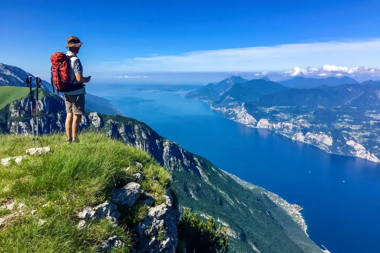 Escursionista con zaino rosso ammira il panorama mozzafiato dal Monte Baldo, con il lago azzurro e le montagne sullo sfondo.