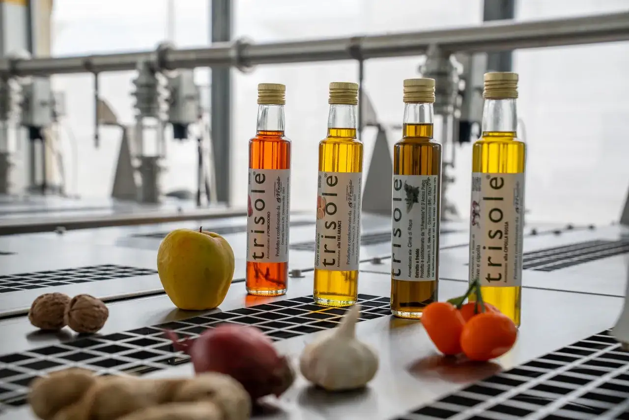 Quattro bottiglie di olio extravergine di oliva aromatizzato, con accanto ingredienti freschi come mela, noci, cipolla e pomodorini.