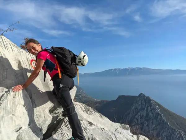 Donna sorridente su roccia con zaino e casco, vista lago e montagne. Un facile sentiero sul monte Pizzocolo.