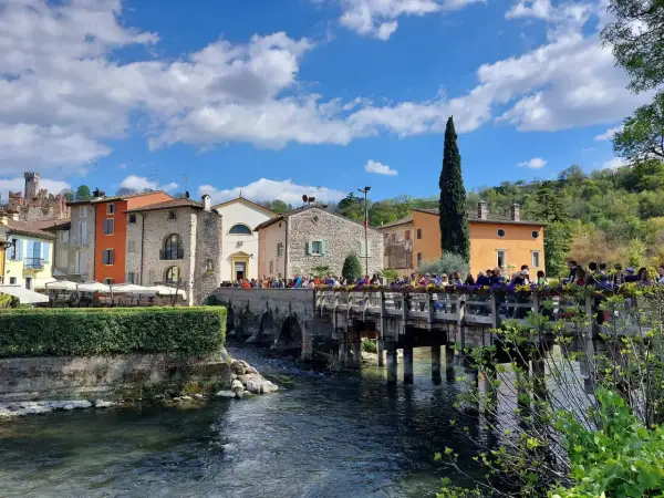 Un pittoresco borghetto mantovano con case colorate, un ponte di legno affollato di gente e un fiume che scorre sotto un cielo azzurro con nuvole bianche.