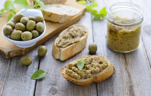 Bruschette con crema di olive, un vasetto di crema di olive e olive verdi su un tagliere di legno.