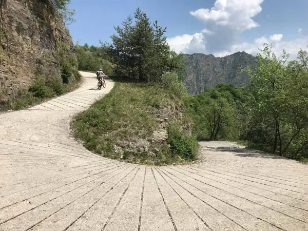 Ciclista su strada sterrata nel Parco Alto Garda Bresciano, tra rocce e vegetazione lussureggiante, con montagne sullo sfondo.