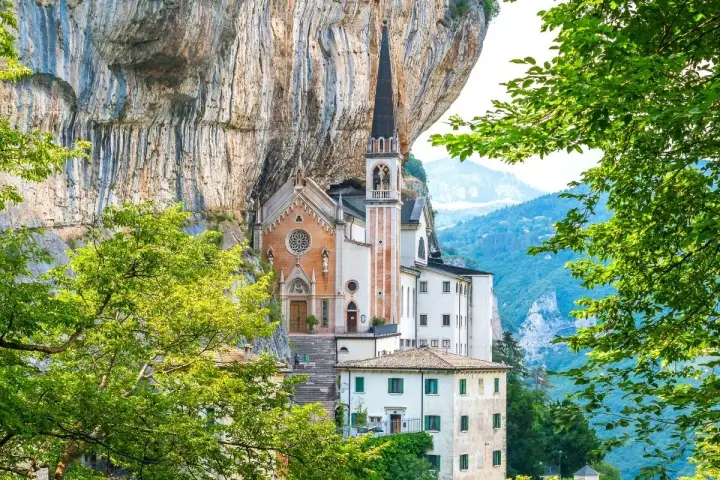 Suggestiva chiesa incastonata nella roccia, un gioiello architettonico vicino al Lago di Garda, circondata da verde lussureggiante e montagne.